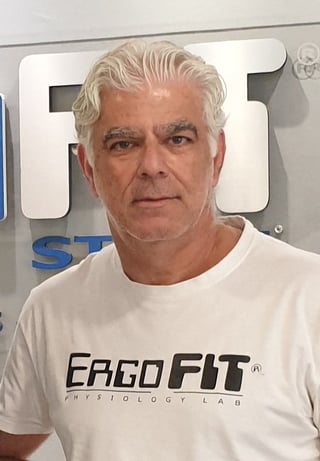 Middle-aged man with gray hair wearing a white ERGO FIT t-shirt, standing in front of signage